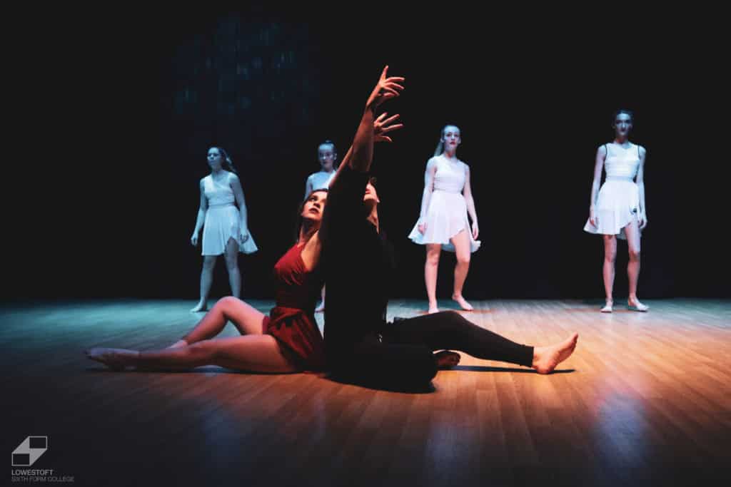 2 performers sitting on floor with hands in air at end of performance. 4 performers standing at the back of the stage.